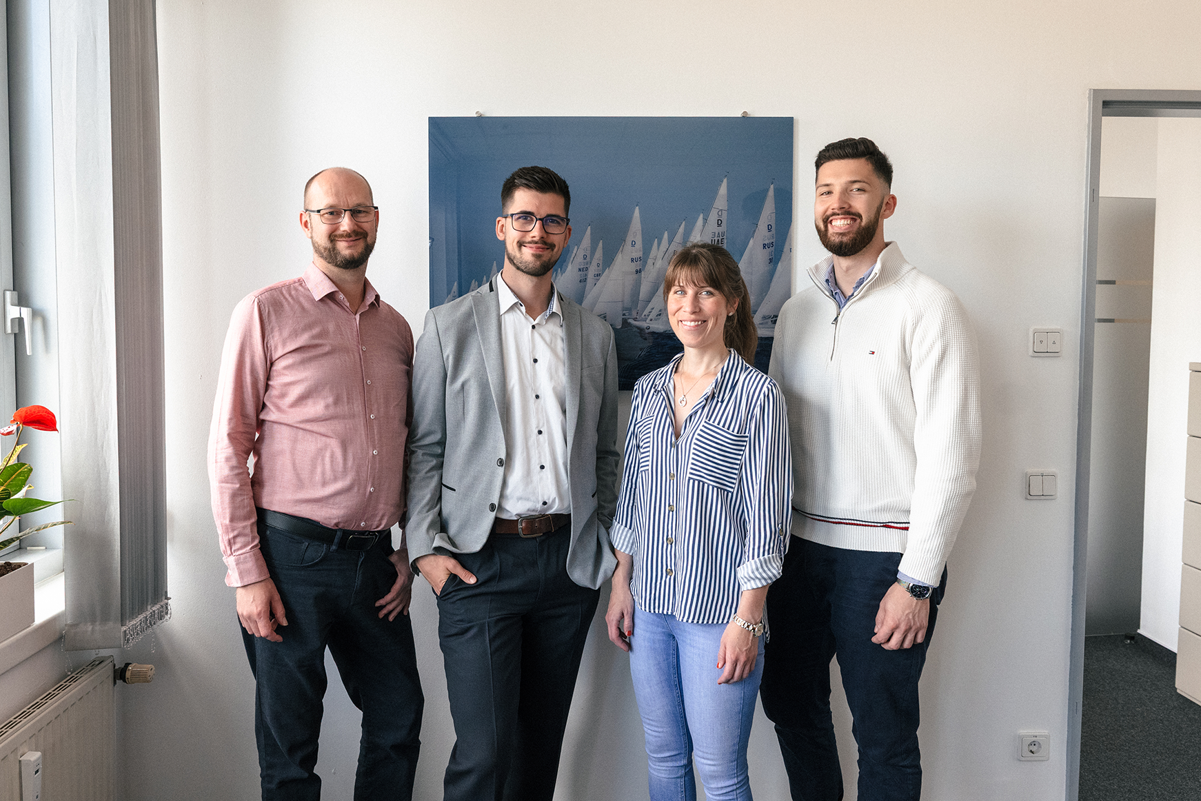 Vier Mitglieder des Provenia-Teams stehen nebeneinander und lächeln in einem Büro vor einem Bild mit Segelbooten.