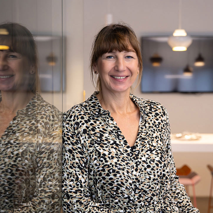 Frau mit gemusterter Bluse steht lächelnd in einem offenen Raum.