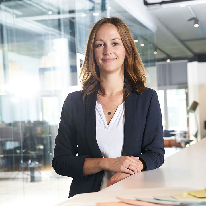 Frau im Blazer steht lächelnd in modernem Glasbüro.