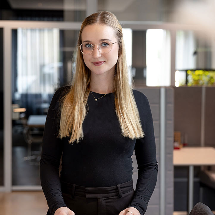 Junge Frau mit Brille steht freundlich in heller Büroumgebung.