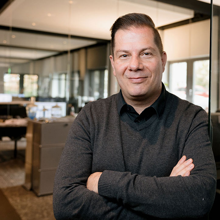 Mann mit verschränkten Armen lächelt in einem offenen Bürobereich.