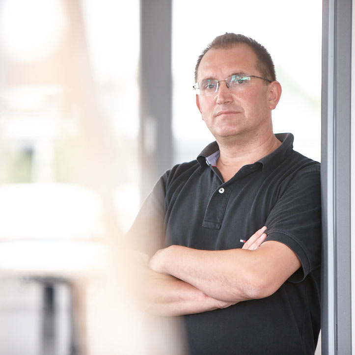 Mann mit verschränkten Armen steht vor Fensterfront.