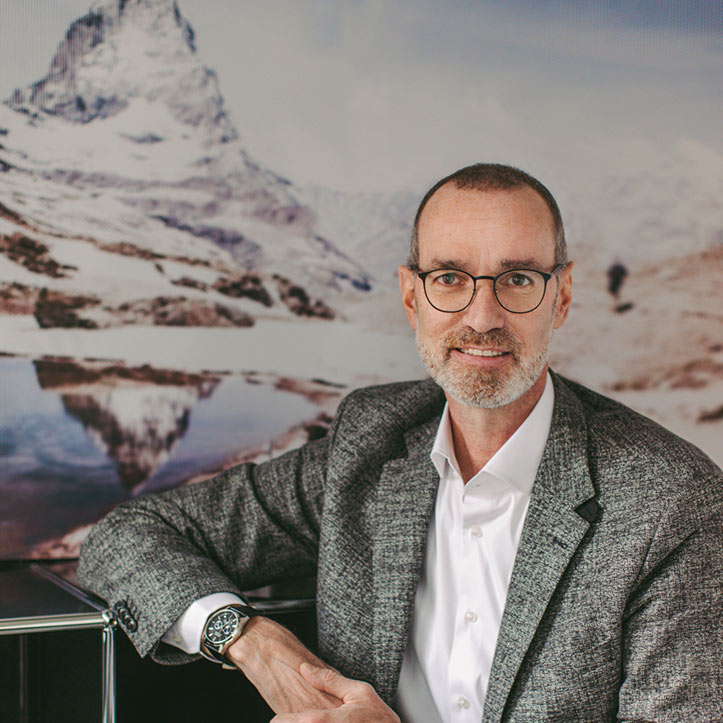 Mann mit Brille und Bart sitzt vor einem Bergmotiv-Hintergrund.