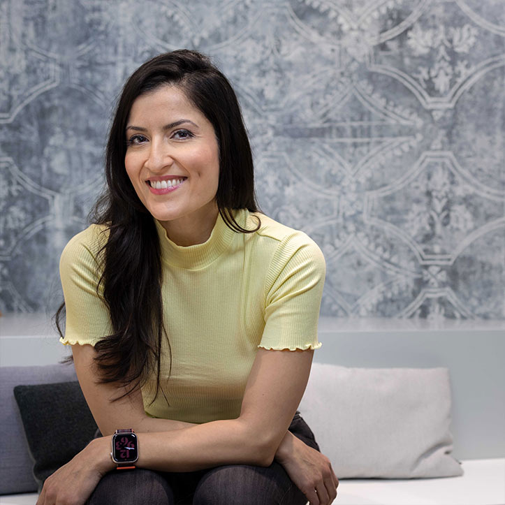 Frau in gelbem Oberteil sitzt lächelnd vor grauer Wand.

