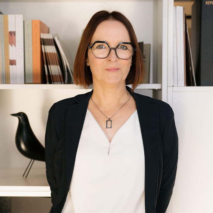 Frau mit Brille und Blazer steht in modernem Arbeitsumfeld.