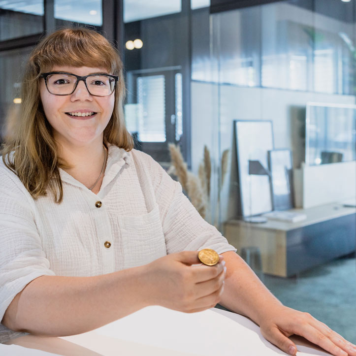 Lächelnde Frau in heller Bluse sitzt in einem offenen Büro.