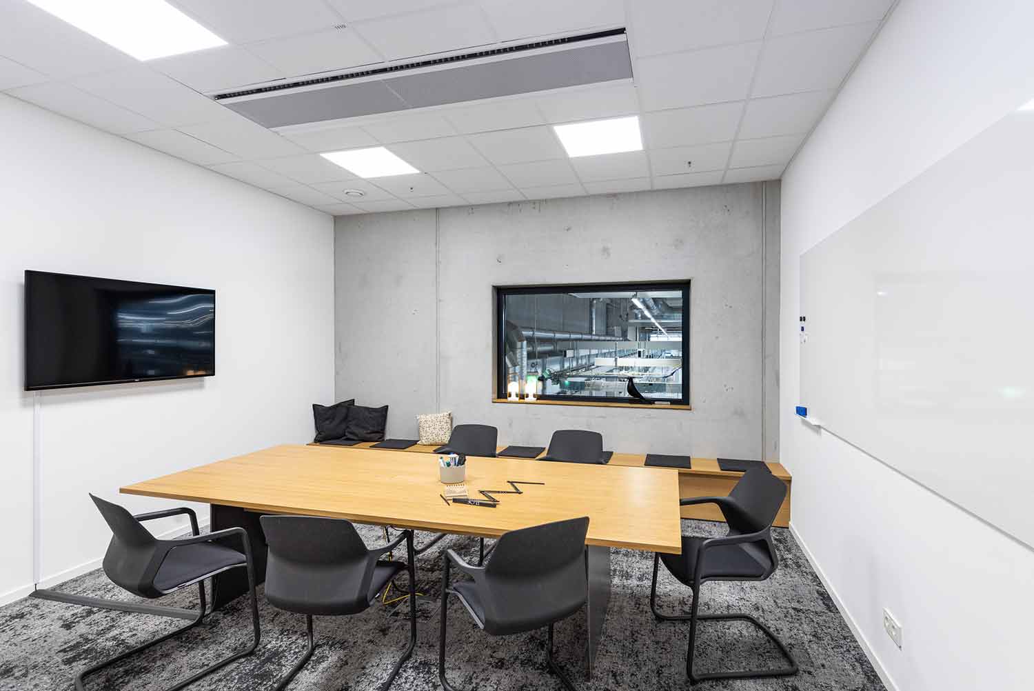 Schlichter Besprechungsraum mit Holztisch, Monitor und grauen Stühlen.