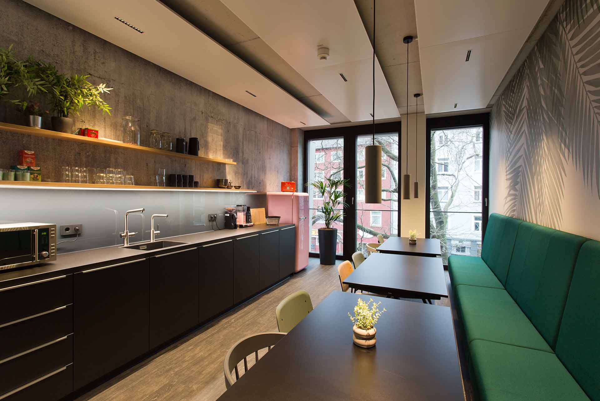 Moderne Küche und Essbereich mit dunklen Schränken, grüner Sitzbank, schwarzen Tischen, Pendelleuchten und großen Fenstern mit Blick auf Bäume und Gebäude.