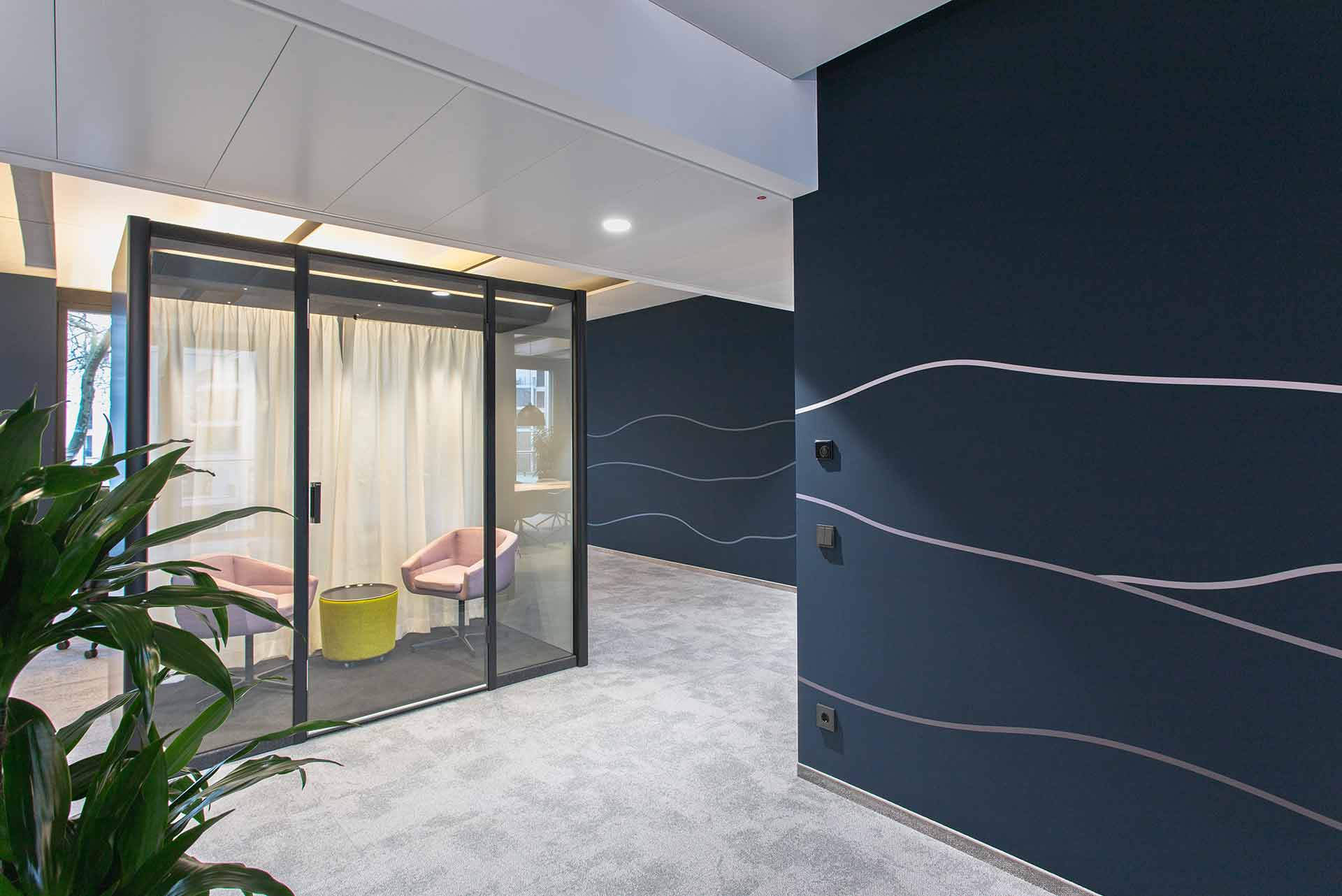 Moderner Büroraum mit dunkelblauen Wänden und geschwungenen silbernen Linien, grauem Teppichboden, einem verglasten Besprechungsbereich mit zwei rosa Stühlen und einem gelben Beistelltisch sowie einer grünen Pflanze im Vordergrund.