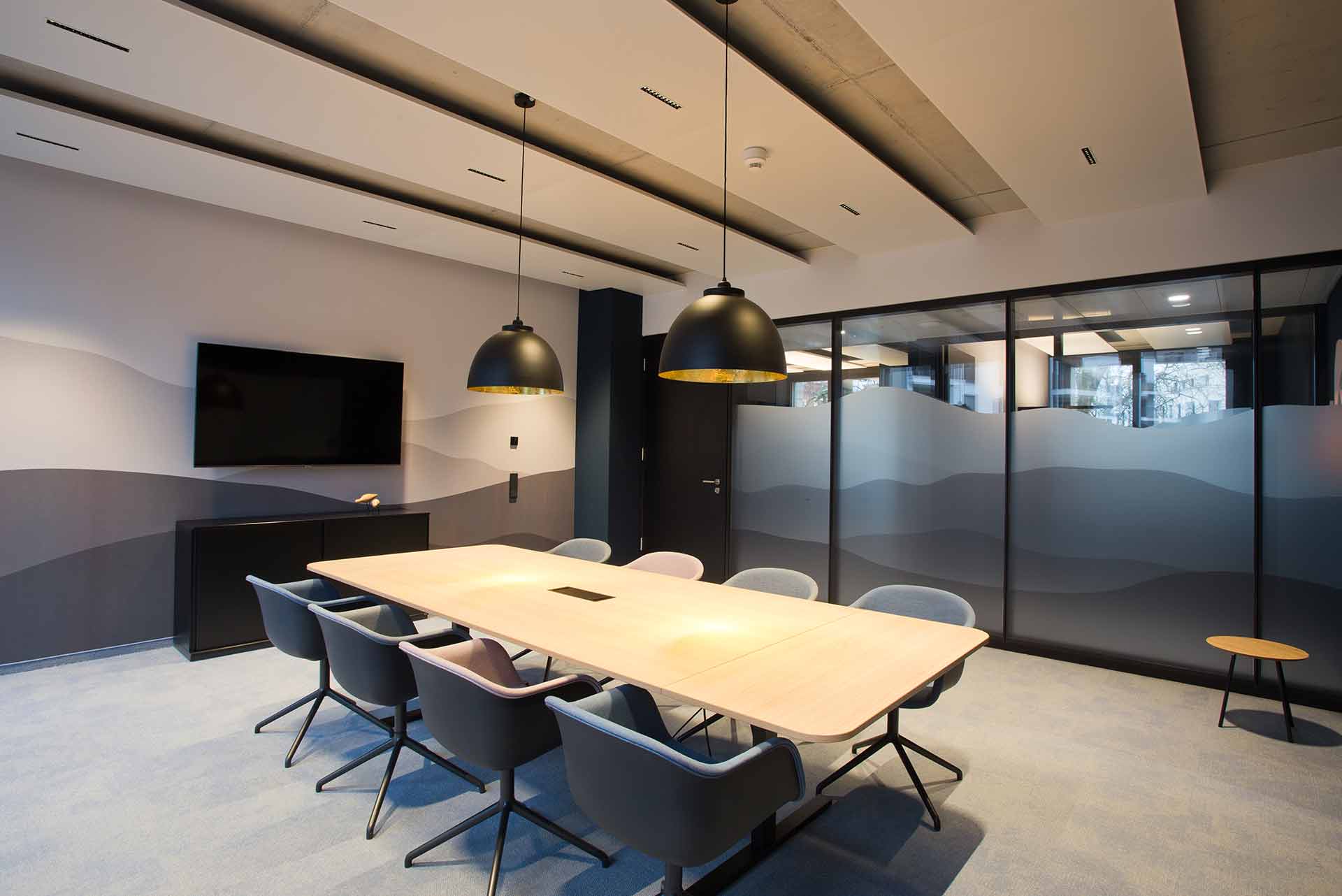 Moderner Konferenzraum mit einem rechteckigen Tisch aus hellem Holz, umgeben von acht schwarz-grau gepolsterten Stühlen, zwei großen schwarzen Pendelleuchten, einem wandmontierten Fernseher und Milchglas-Trennwänden mit Wellenmustern.