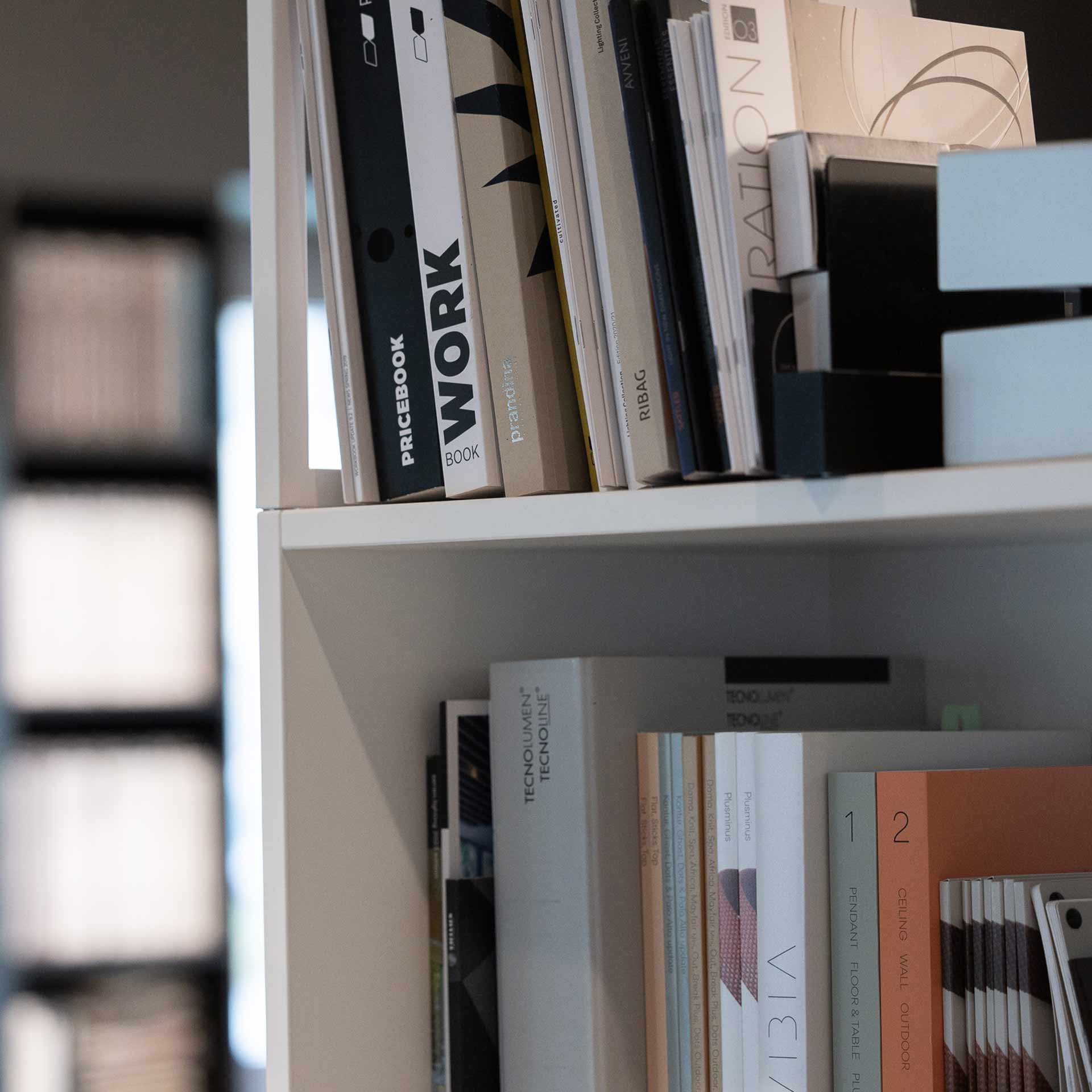 Close-up of books and catalogs organized on white shelves with visible titles like WORK, TECNOLOUMEN TECNOLINE, and numbered design sections.