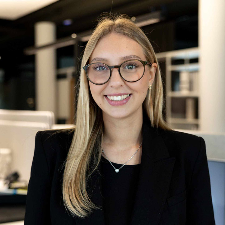 Junge Frau mit Brille und blondem Haar lächelt freundlich in einem modernen Büro.