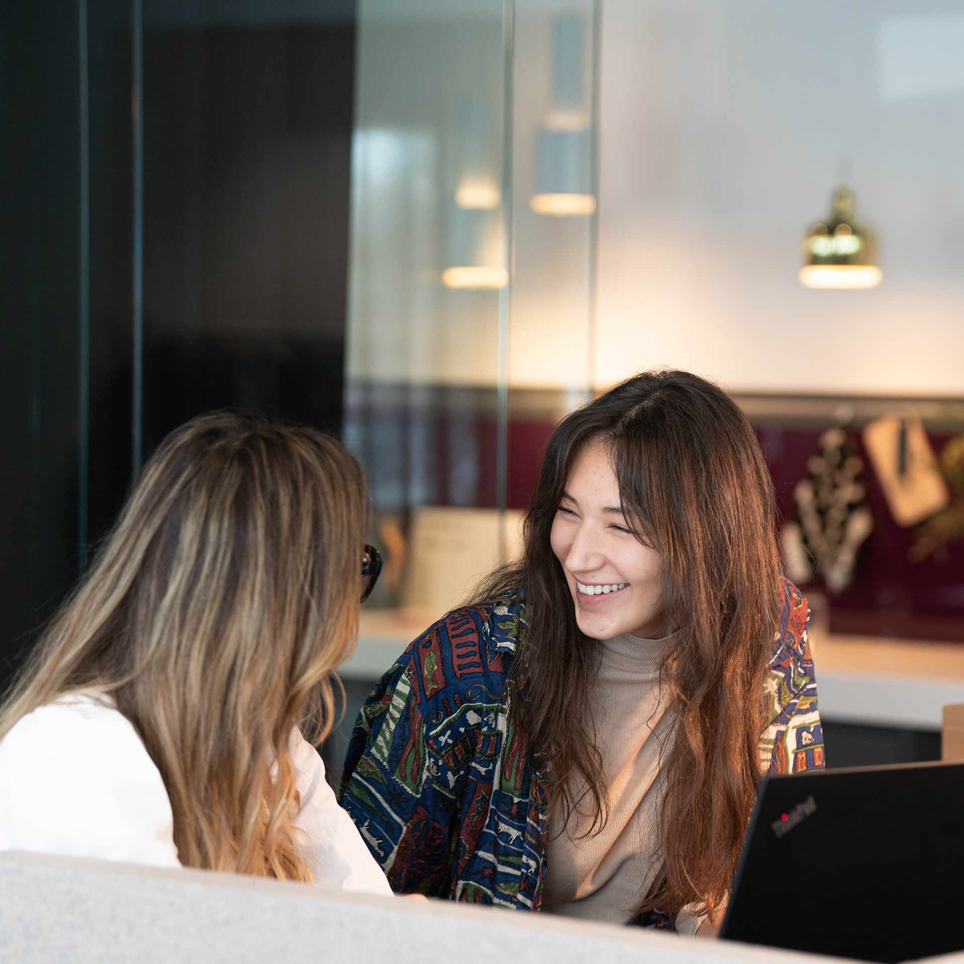 Zwei Kolleginnen unterhalten sich lächelnd in einem modernen Büro.