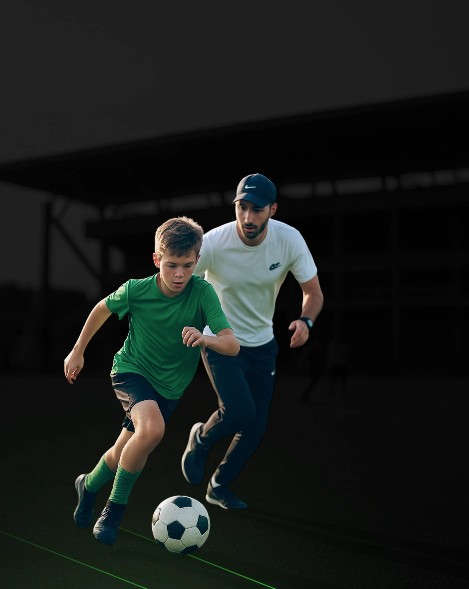 Ragazzo in maglietta verde e calzoncini neri dribbla un pallone da calcio mentre un uomo in maglietta bianca e cappellino lo segue durante un allenamento.