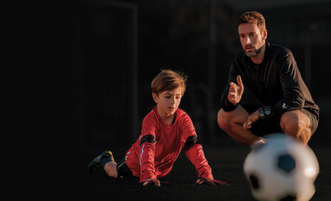 Bambino in maglia rossa con guanti da portiere che si esercita a terra mentre un allenatore in abbigliamento sportivo nero gli dà istruzioni vicino a un pallone da calcio sfocato in primo piano.