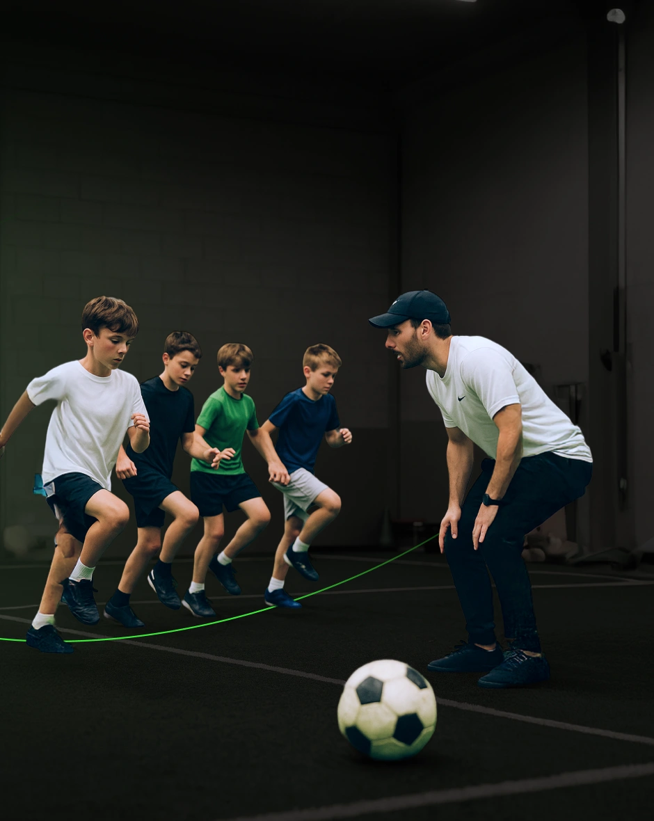 Allenatore con berretto che osserva cinque ragazzi mentre si esercitano su un campo da calcio indoor.