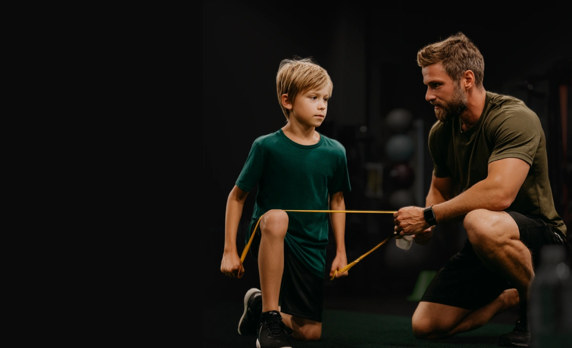 Bambino con maglietta verde che esegue esercizio con elastico mentre un uomo lo assiste in palestra.