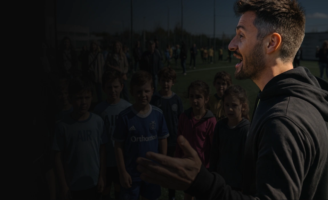 Allenatore con felpa nera che parla con un gruppo di bambini su un campo da calcio all'aperto.