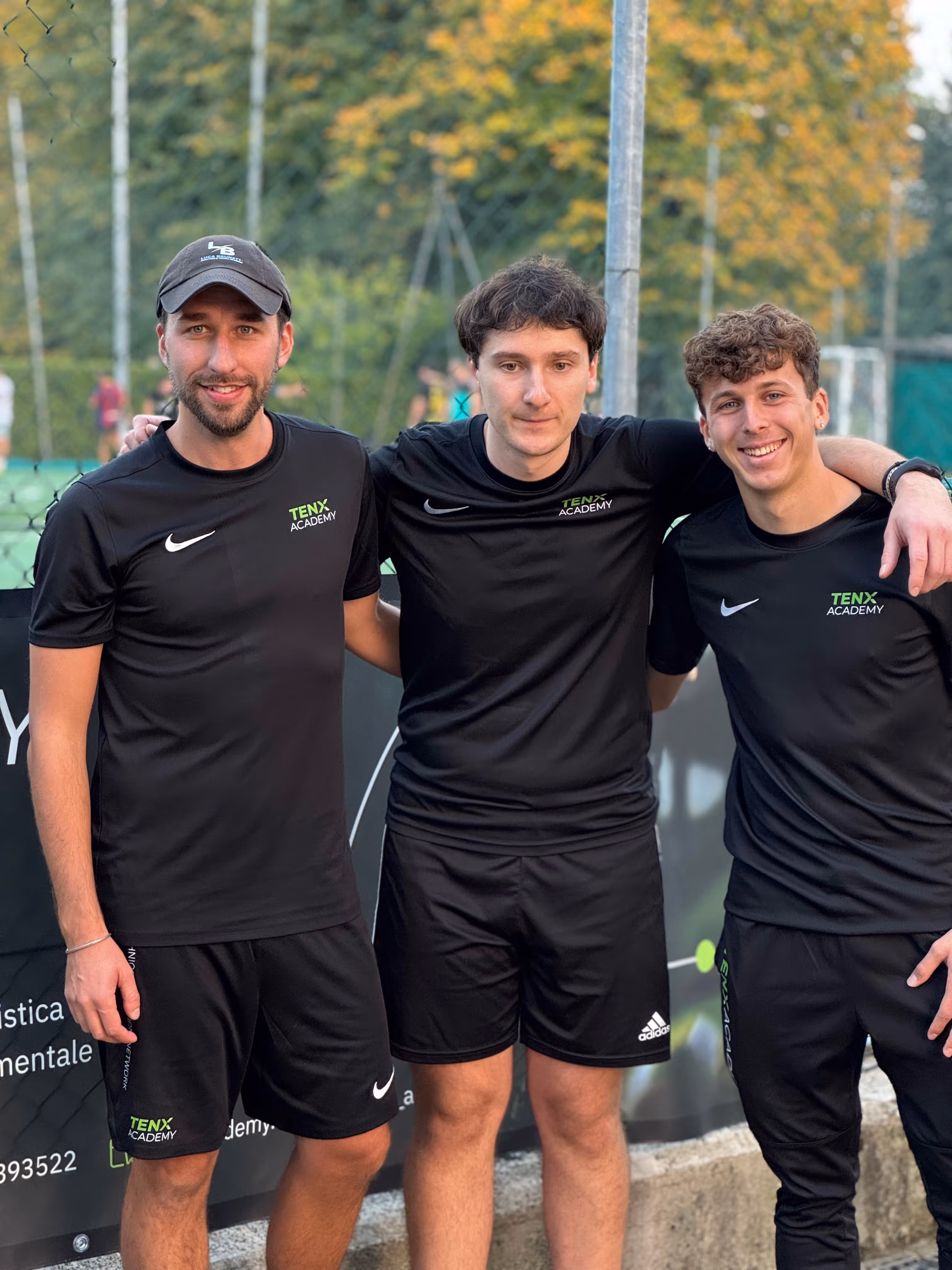 Tre giovani uomini in abbigliamento sportivo nero con logo TenX Academy sorridono all'aperto vicino a un campo da tennis.