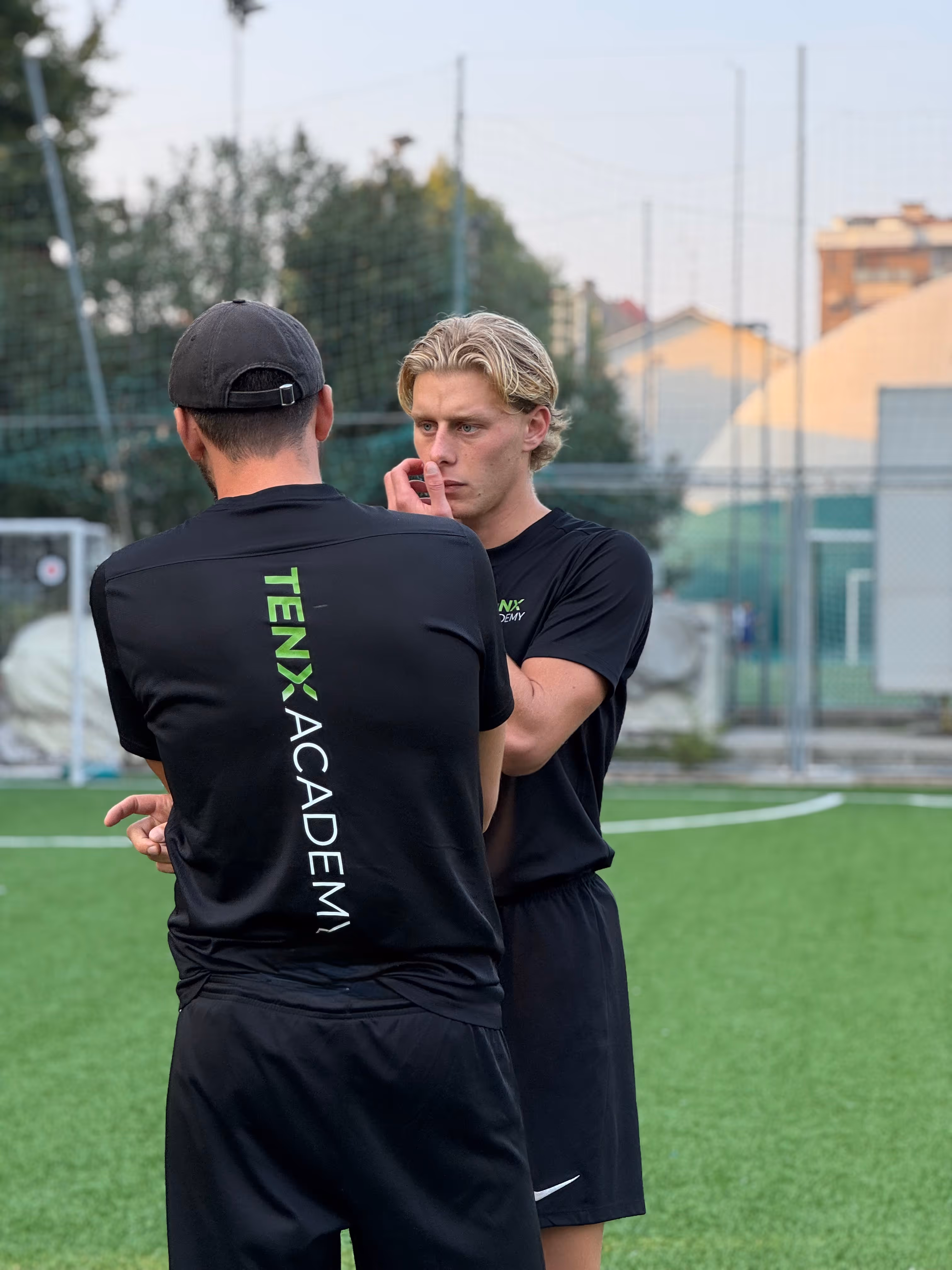 Due uomini in divise nere della TenX Academy conversano su un campo da calcio in erba sintetica.