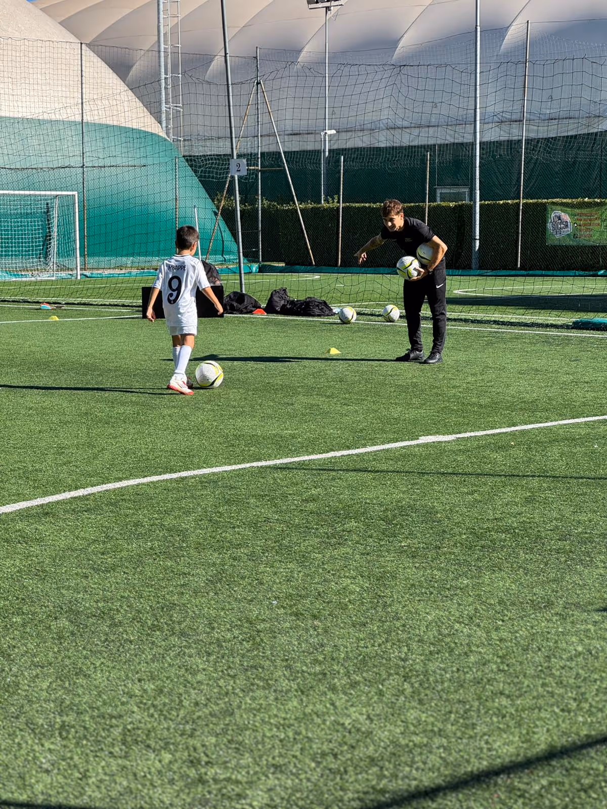 Bambino con maglia numero 9 che calcia un pallone da calcio durante l'allenamento, con allenatore che tiene due palloni su un campo di calcio in erba sintetica.