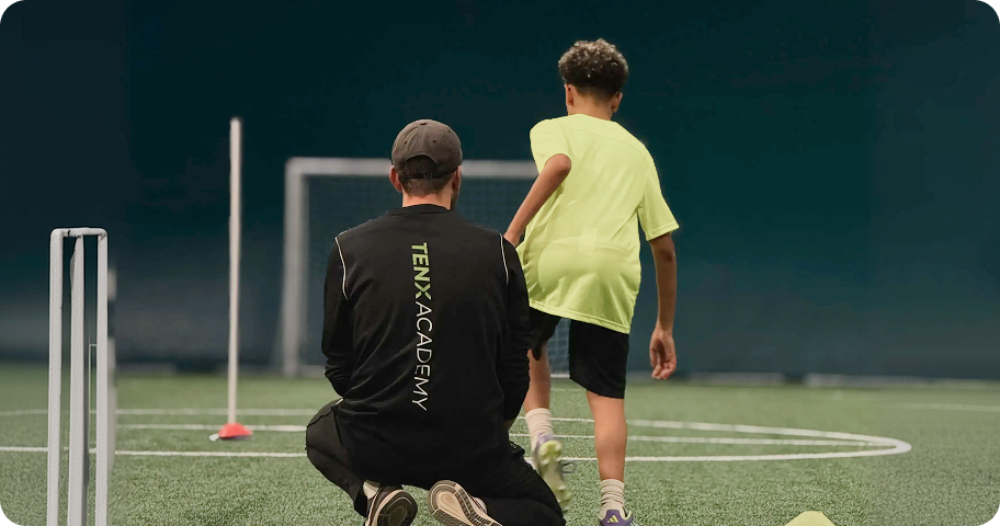 Allenatore in felpa nera con logo TENX ACADEMY osserva ragazzo in maglietta gialla mentre calcia un pallone su campo da calcio.
