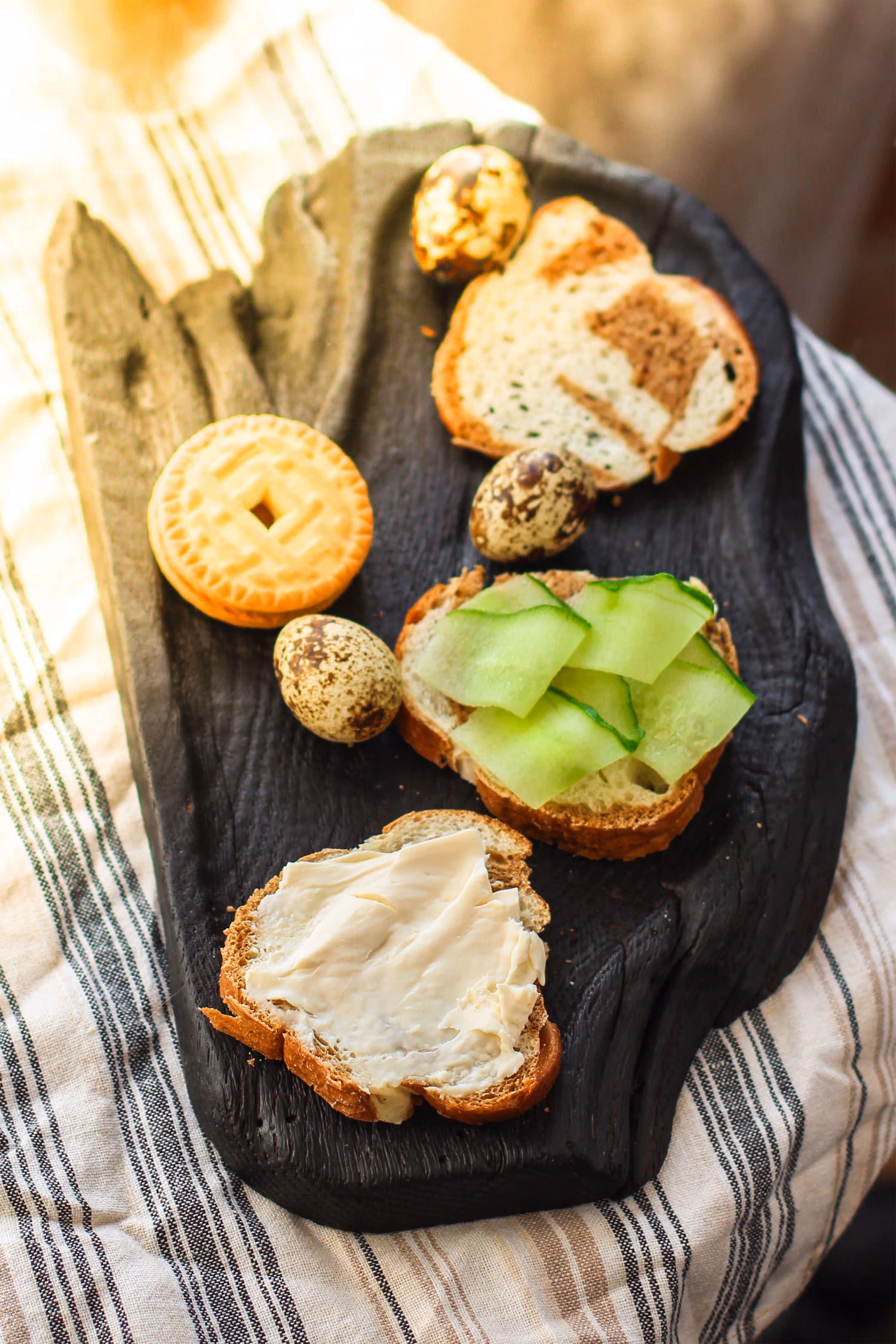Verschiedene Häppchen auf einem dunklen Holzbrett, darunter Gurkenscheiben auf Brot, Brot mit Butter, kleine Eier und ein runder Keks.