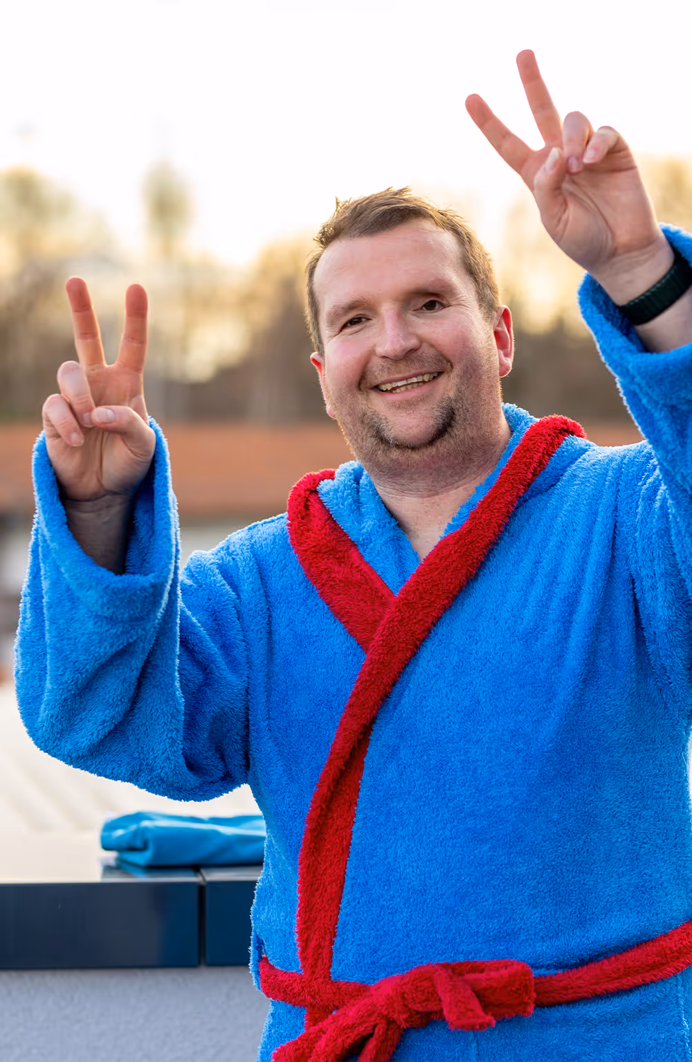 Mann in blauem Bademantel mit roten Akzenten macht mit beiden Händen das Victory-Zeichen und lächelt.