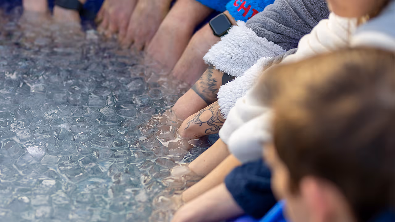Mehrere Hände tauchen in einen Behälter mit Eiswasser ein, einige mit Uhren und Tätowierungen sichtbar.