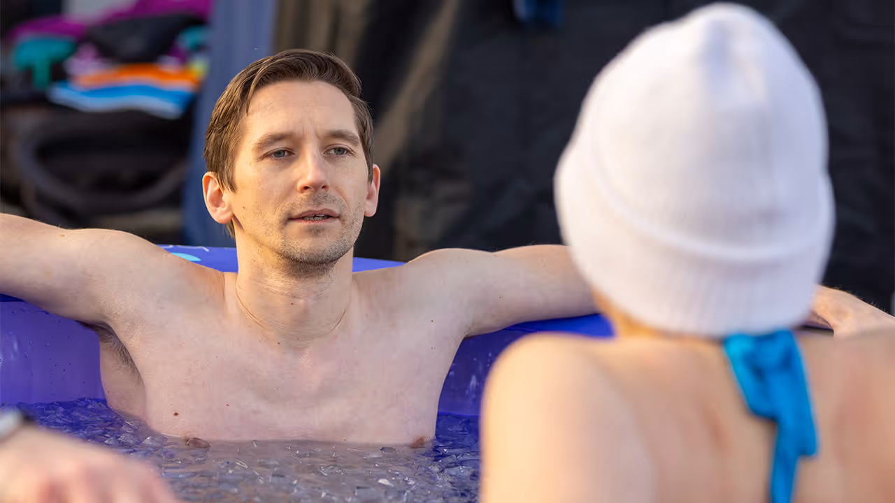 Mann mit nacktem Oberkörper entspannt in einem mit Eis gefüllten Behälter, während eine Person mit weißer Mütze und blauem Badeanzug davor sitzt.