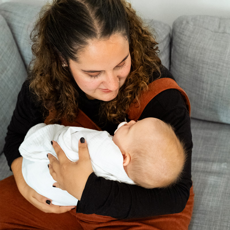 Woman with curly hair gently holding and looking down at a sleeping baby wrapped in a white blanket on a gray couch.