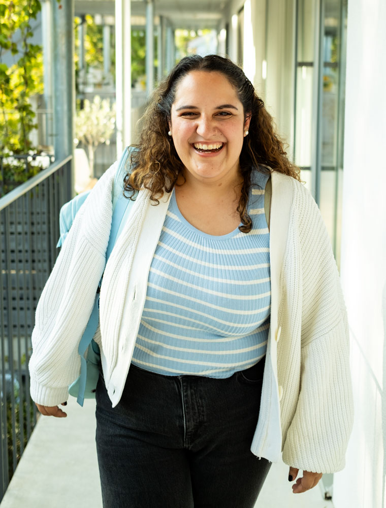 Smiling woman with curly hair wearing a white cardigan, blue striped top, and black pants walking outdoors with a light blue backpack.