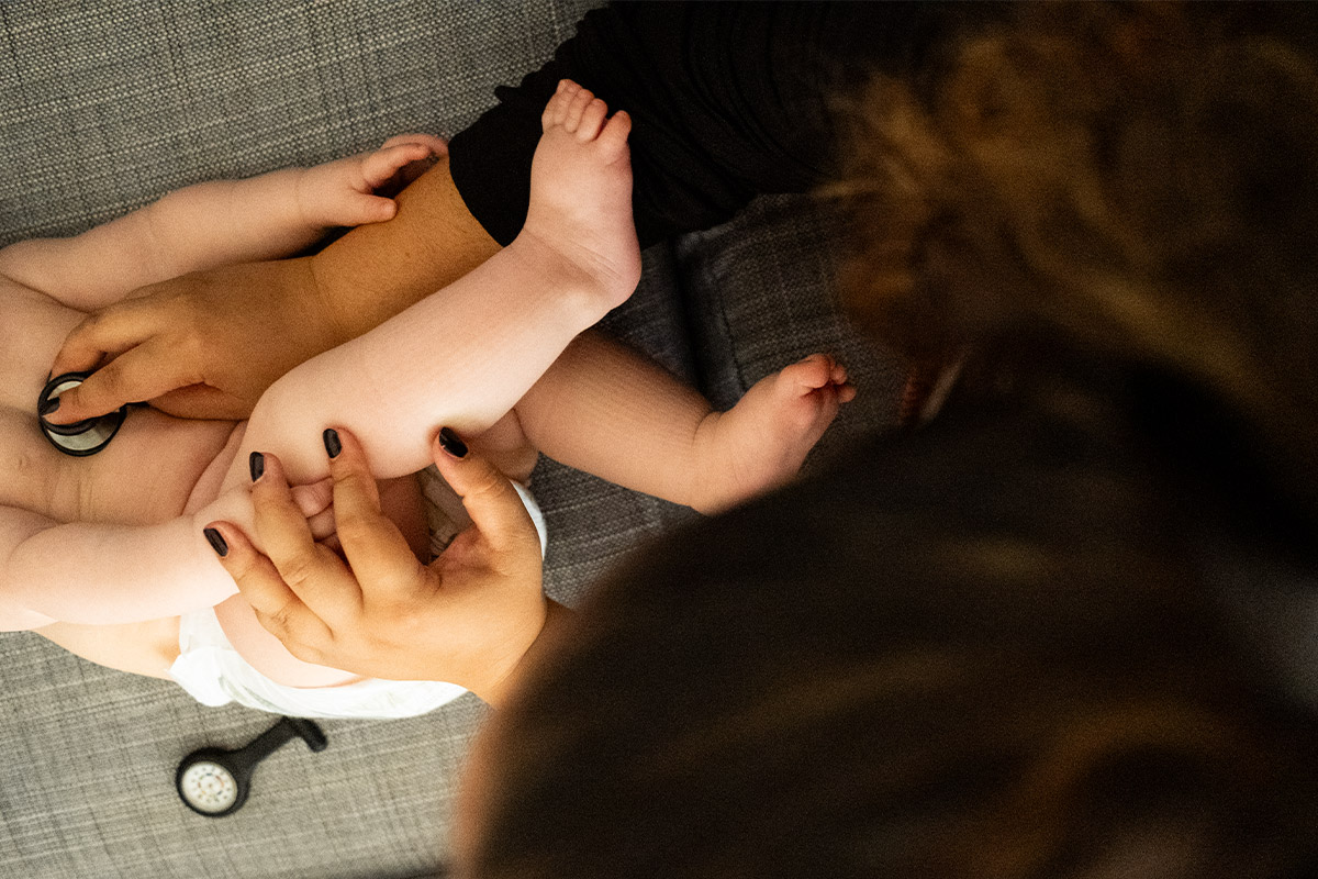 Close-up of a healthcare professional using a stethoscope to examine a baby lying on a gray fabric surface.