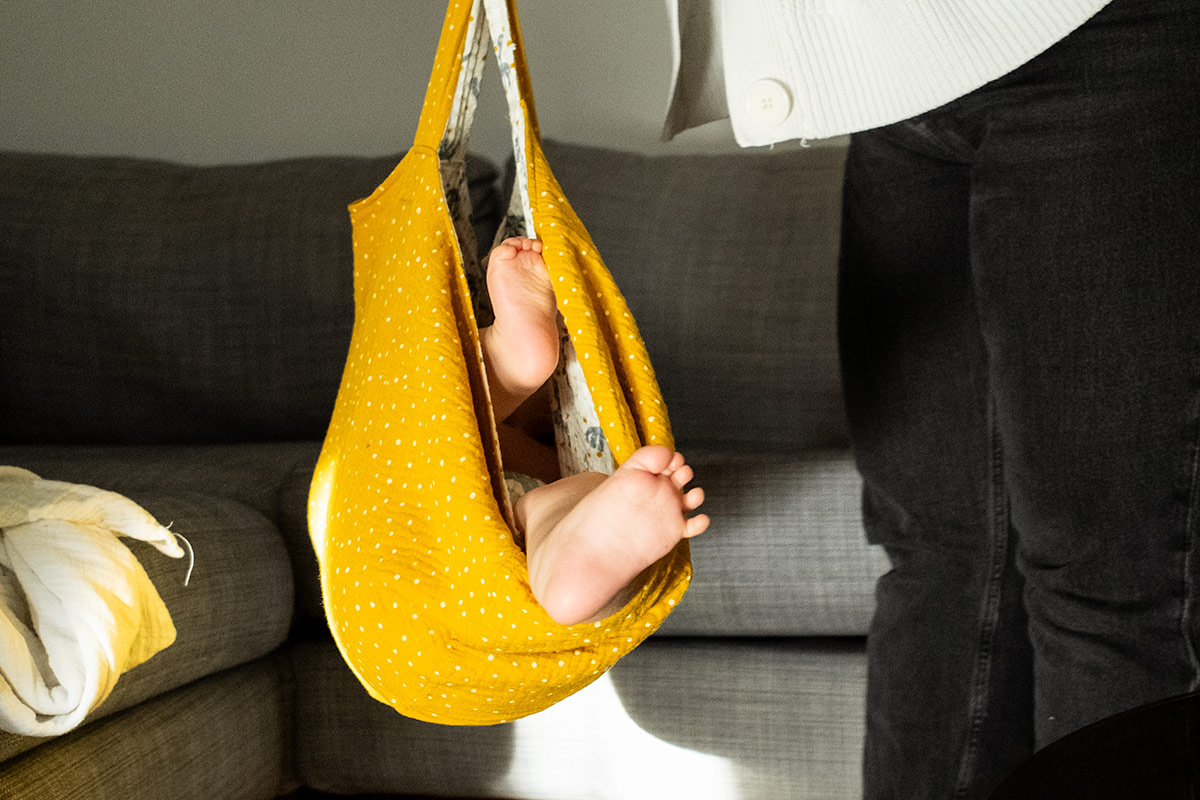 Baby's bare feet visible inside a yellow fabric sling being held by an adult wearing black pants and a white sweater near a gray couch.