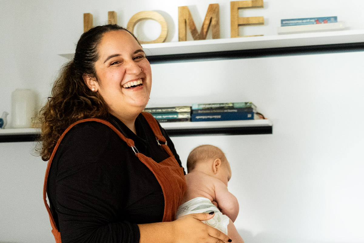 Smiling woman with curly hair holding a baby wearing a diaper against a white wall with shelves and wooden letters spelling HOME.