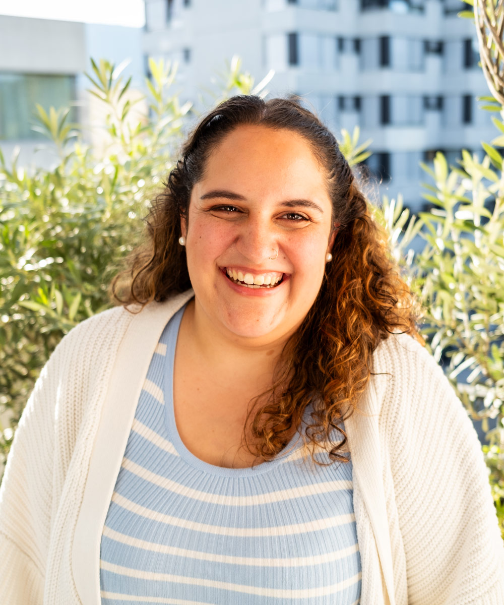 Smiling woman with curly hair wearing a striped blue top and white cardigan outdoors with greenery and buildings in the background.