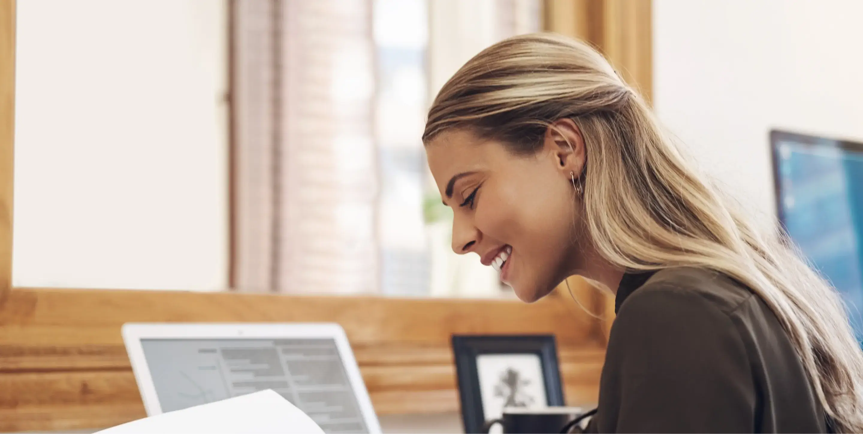 A woman smiles while reading a document at her desk with a laptop nearby.