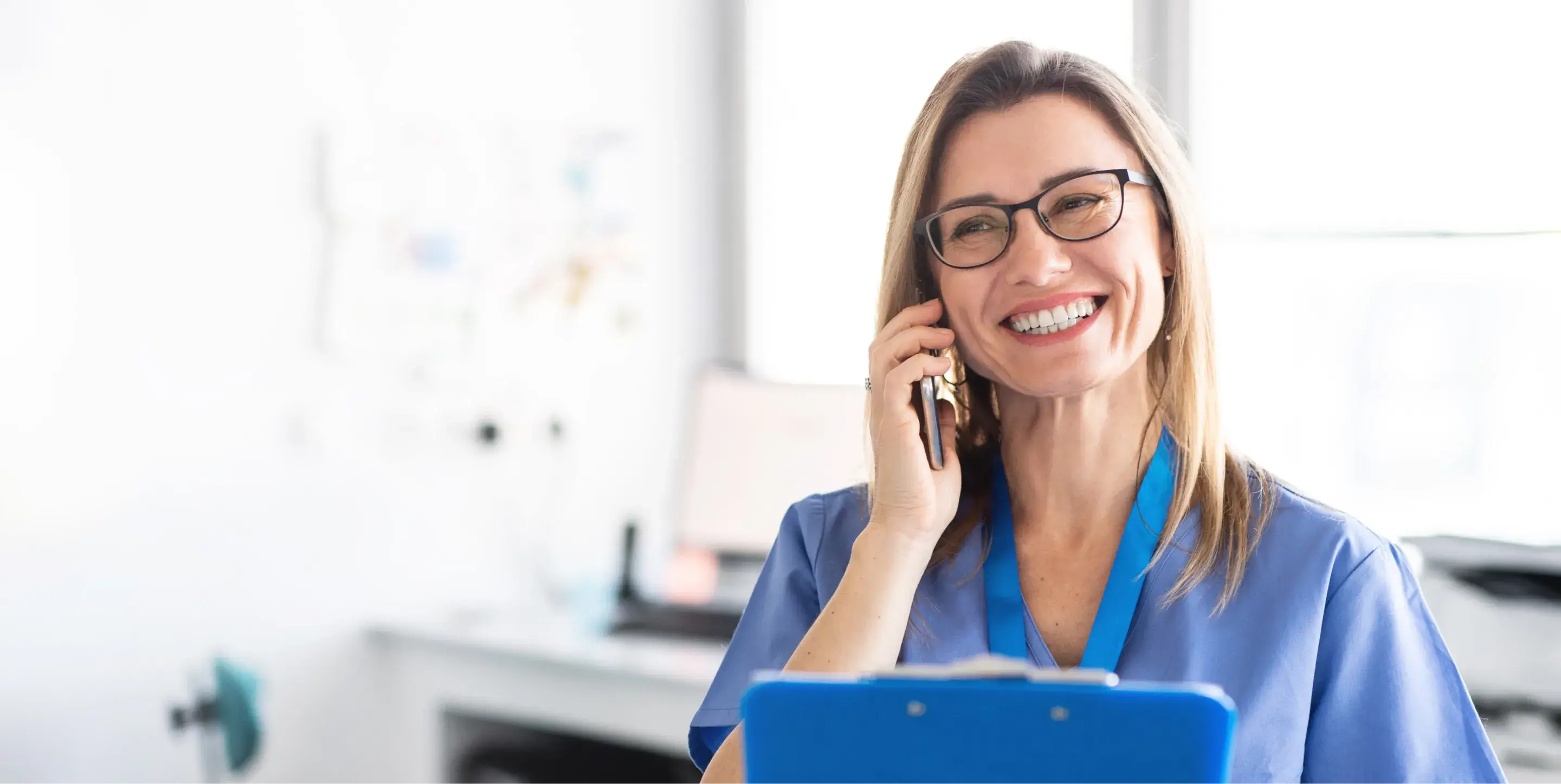 A smiling woman in medical scrubs talks on a phone, holding a clipboard.