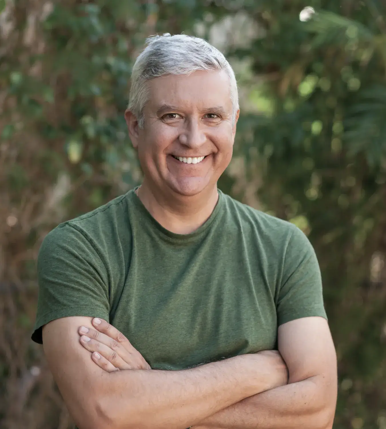 A man with gray hair smiles while standing with arms crossed, wearing a green shirt outdoors.