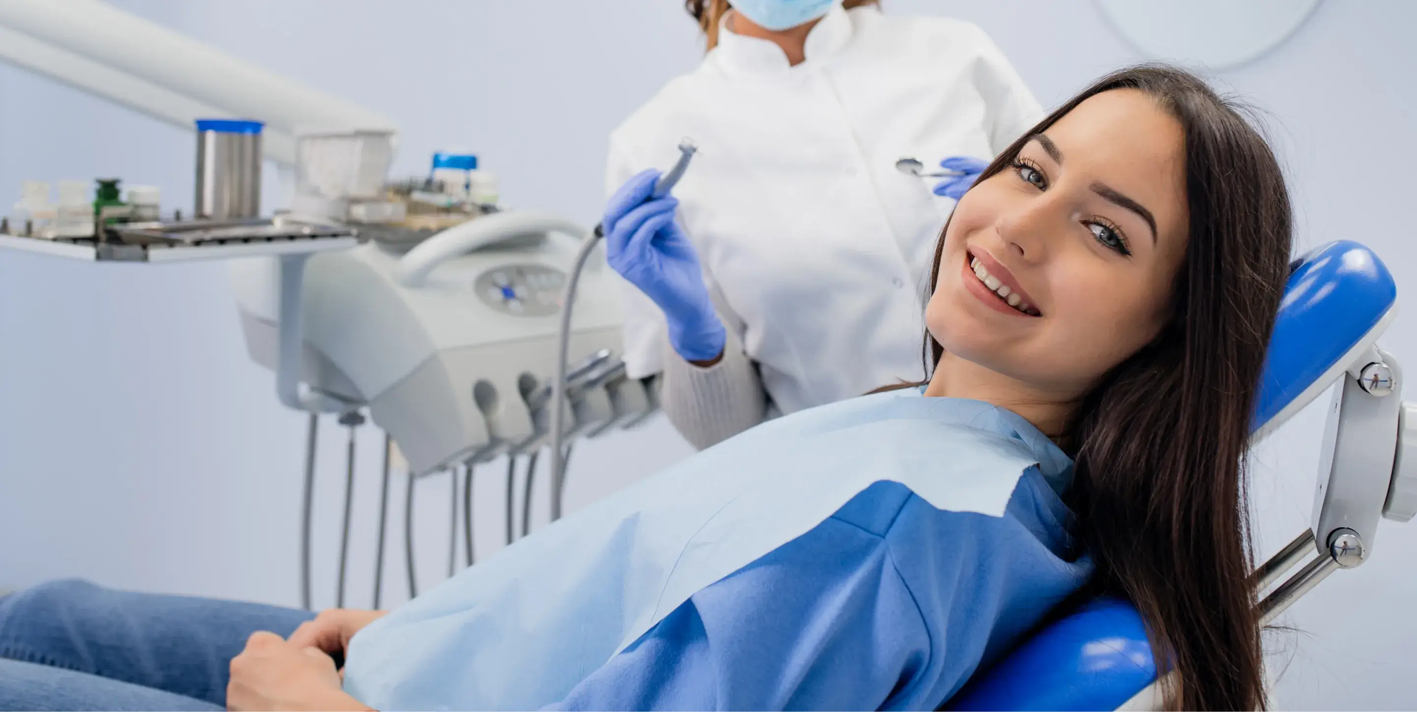 A woman smiles while sitting in a dentist's chair, with a dentist holding dental tools nearby.