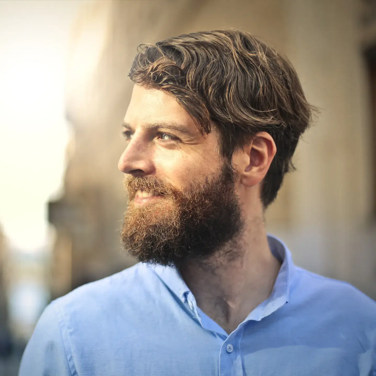 Man with a beard and blue shirt smiling while looking to the side outdoors.