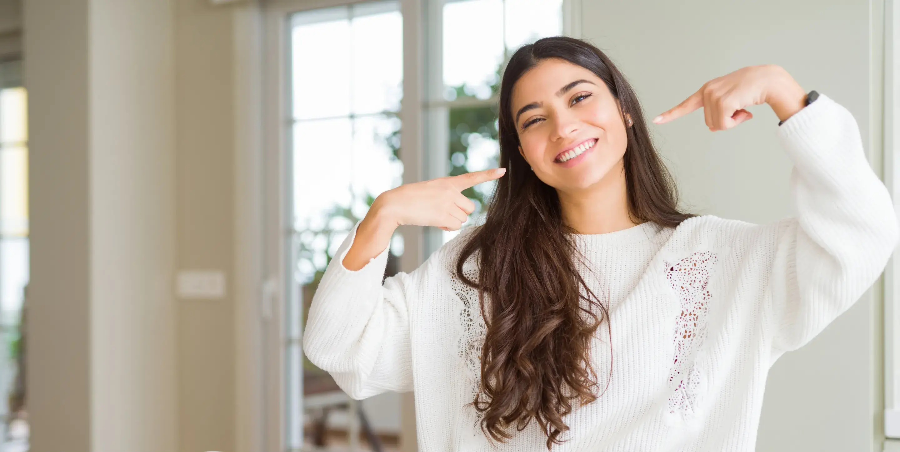 Woman smiling and pointing to her teeth, standing indoors in a white sweater.