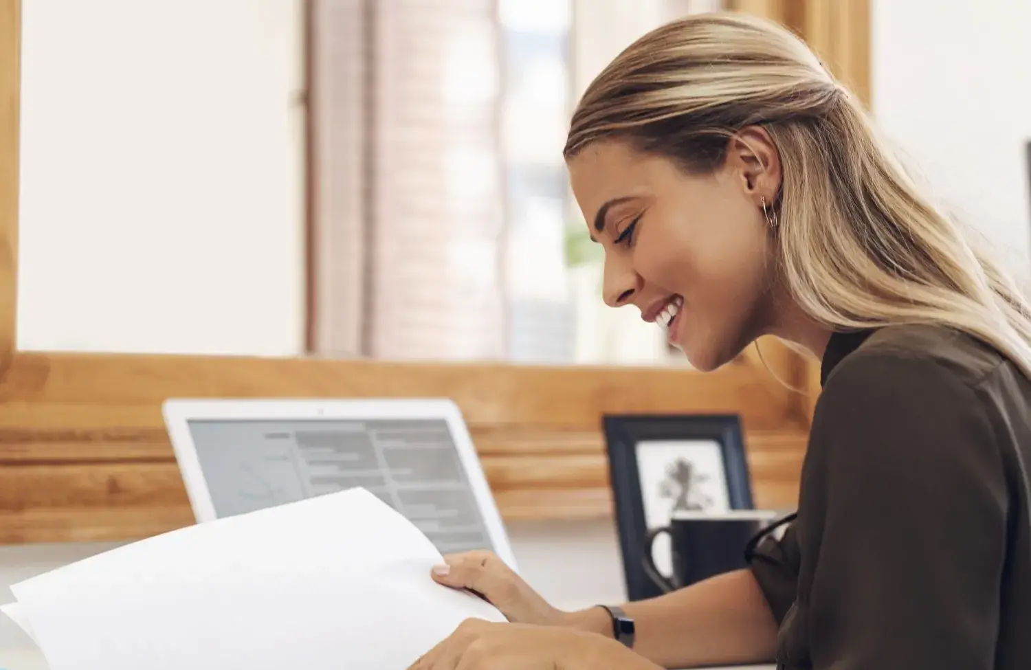 A woman smiles while reading papers at her desk, with a computer and framed picture nearby.