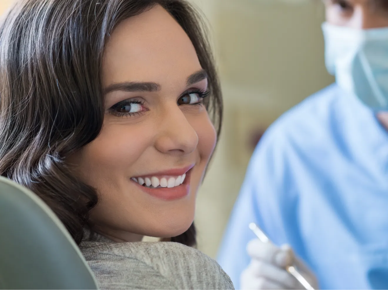 A woman smiles while sitting in a dental chair, with a dentist nearby.
