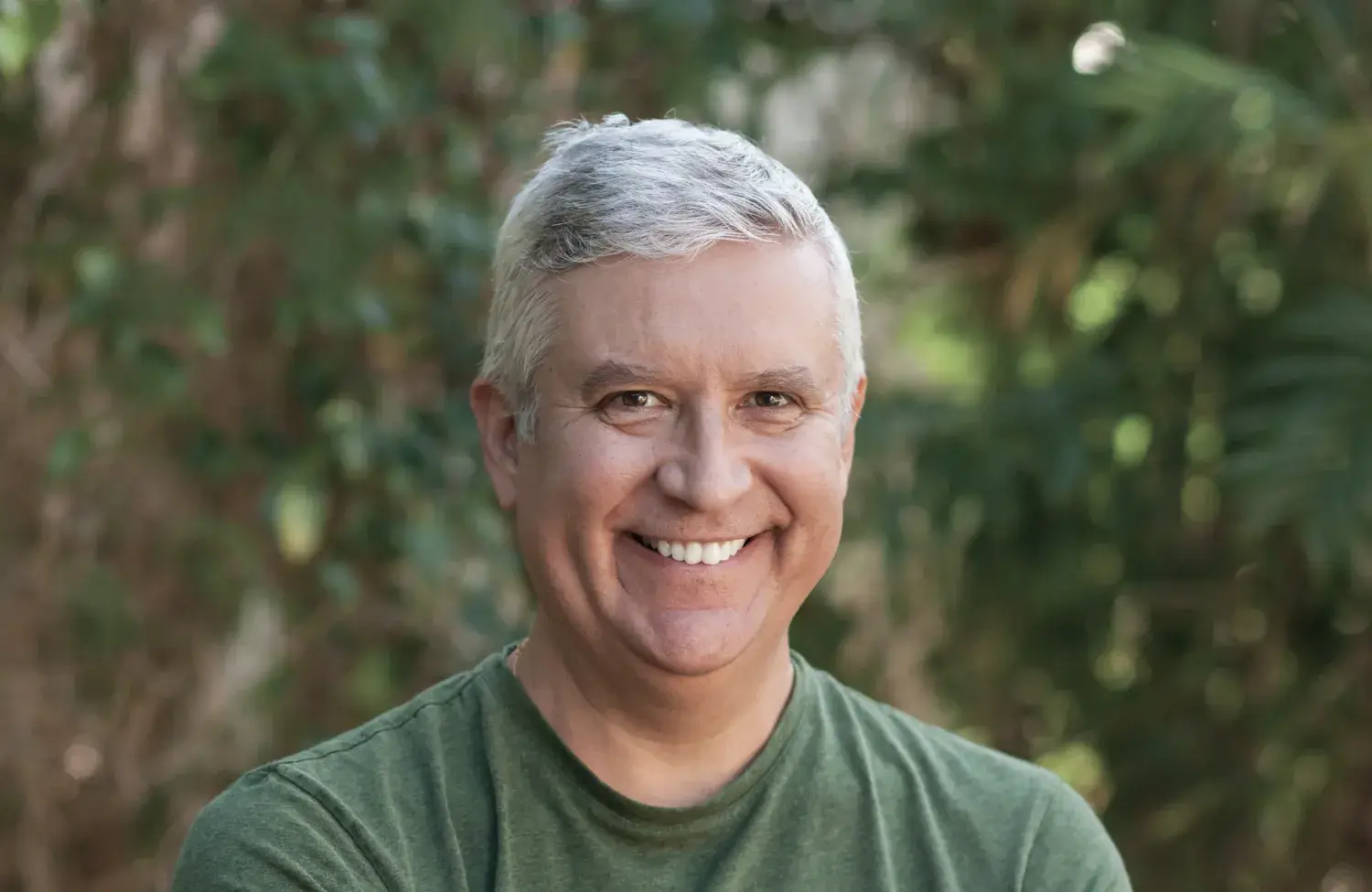 A man with gray hair smiles warmly, standing outdoors with a blurred green background.