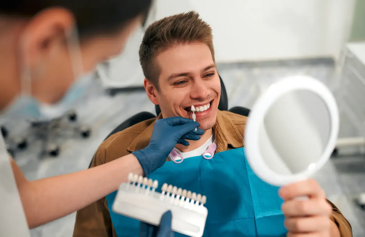 A dentist helps a man choose tooth color using a shade guide while he holds a mirror.