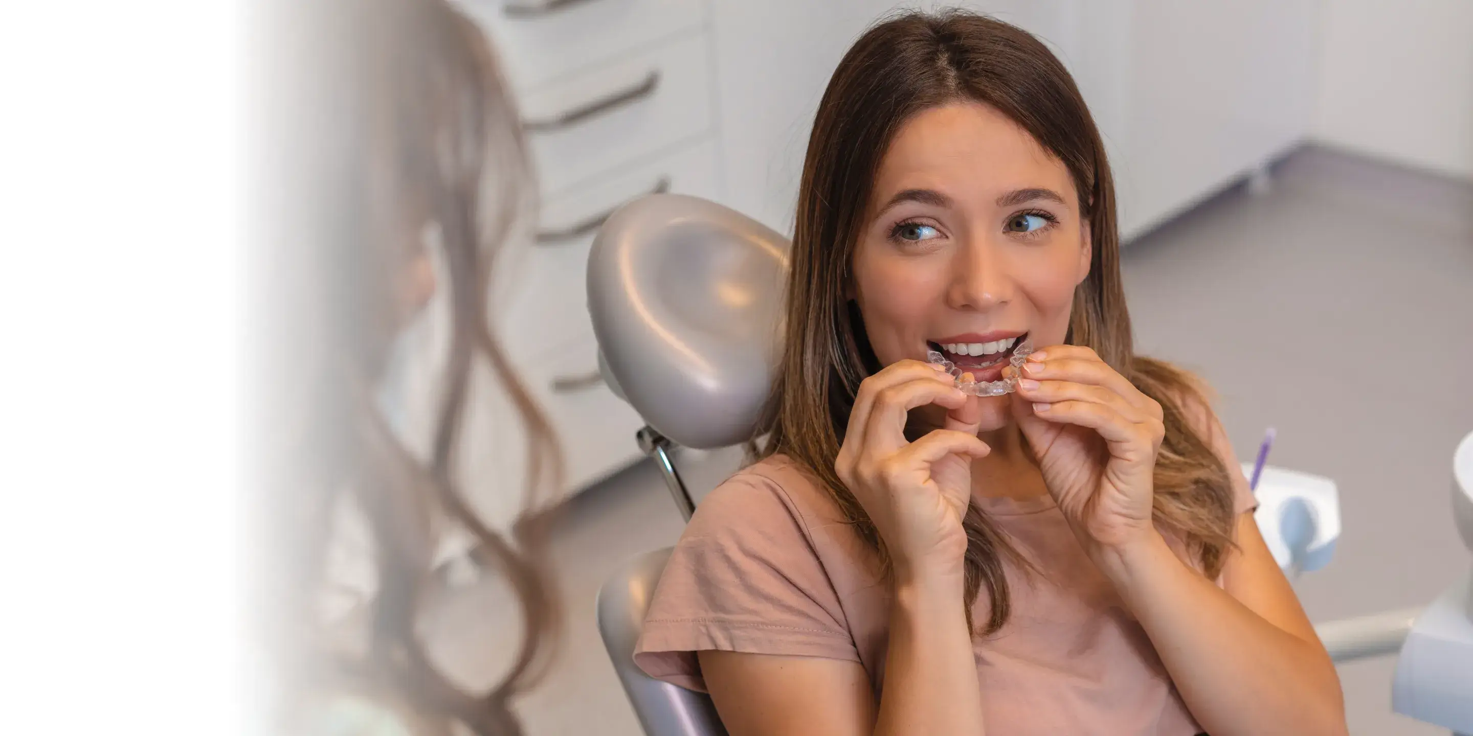 A woman sits in a dental chair, placing a clear aligner over her teeth.