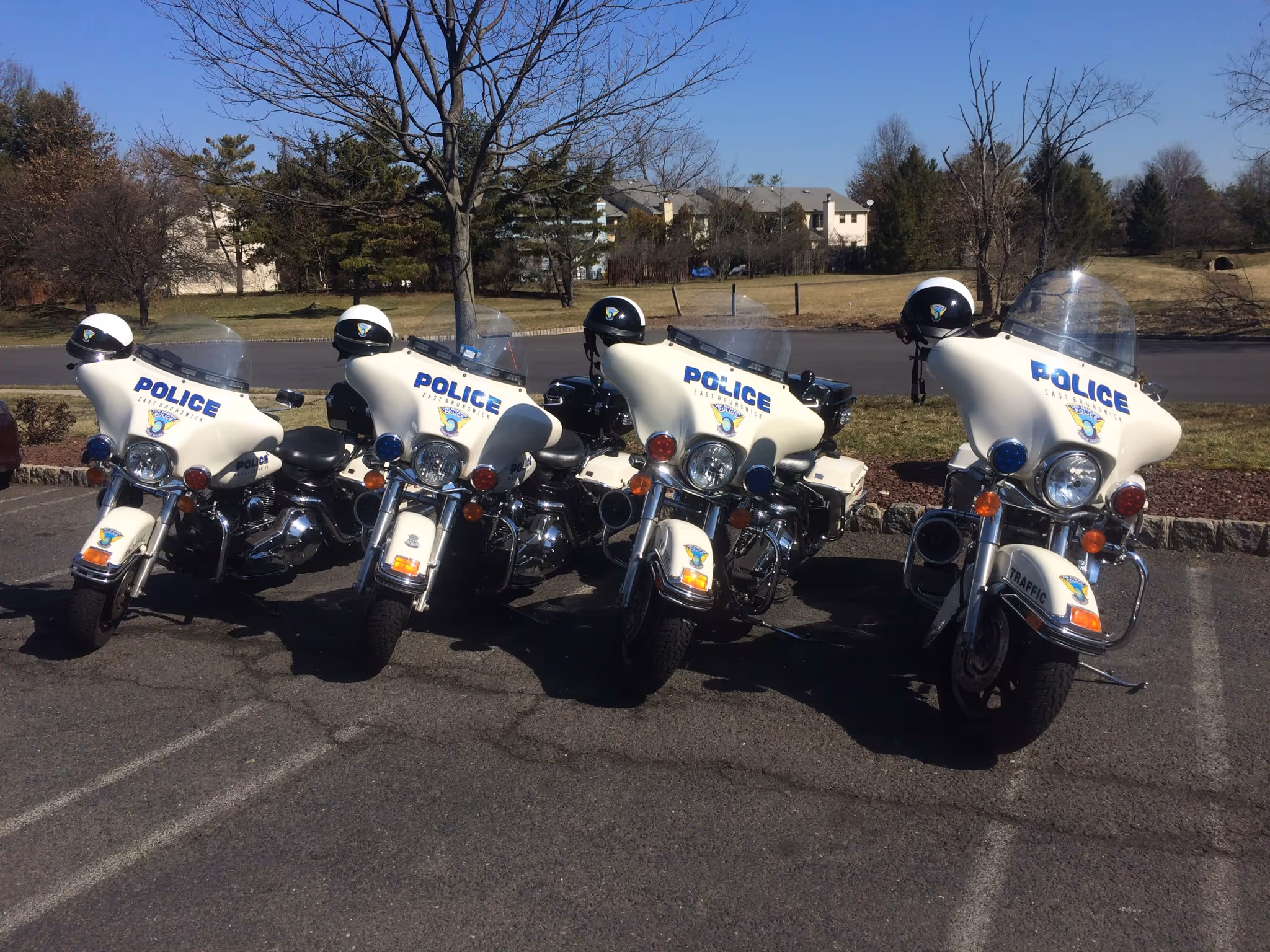 Four white police motorcycles lined up in a parking area, with clear skies and trees in the background.