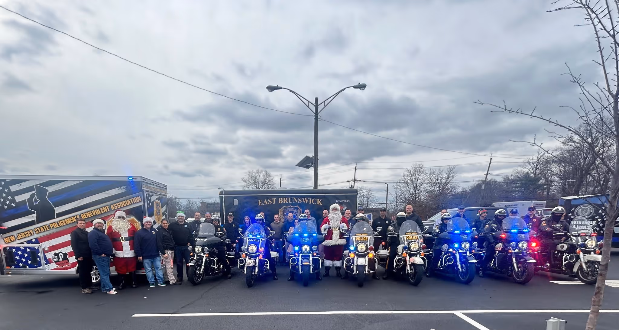 A group of police officers and individuals dressed as Santa Claus posing together in front of emergency vehicles, with police motorcycles lined up and flashing lights on, under a cloudy sky.