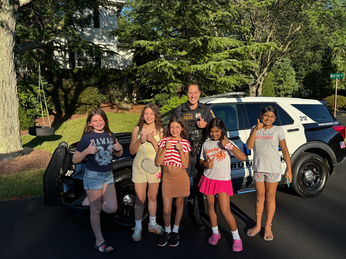 A police officer stands near a patrol car while posing for a photo with six girls outdoors. The children are smiling and giving thumbs up in front of a house with trees.
